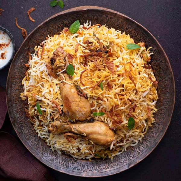 Chicken biryani with two chicken legs, fried onions, and mint in an ornate metal bowl.